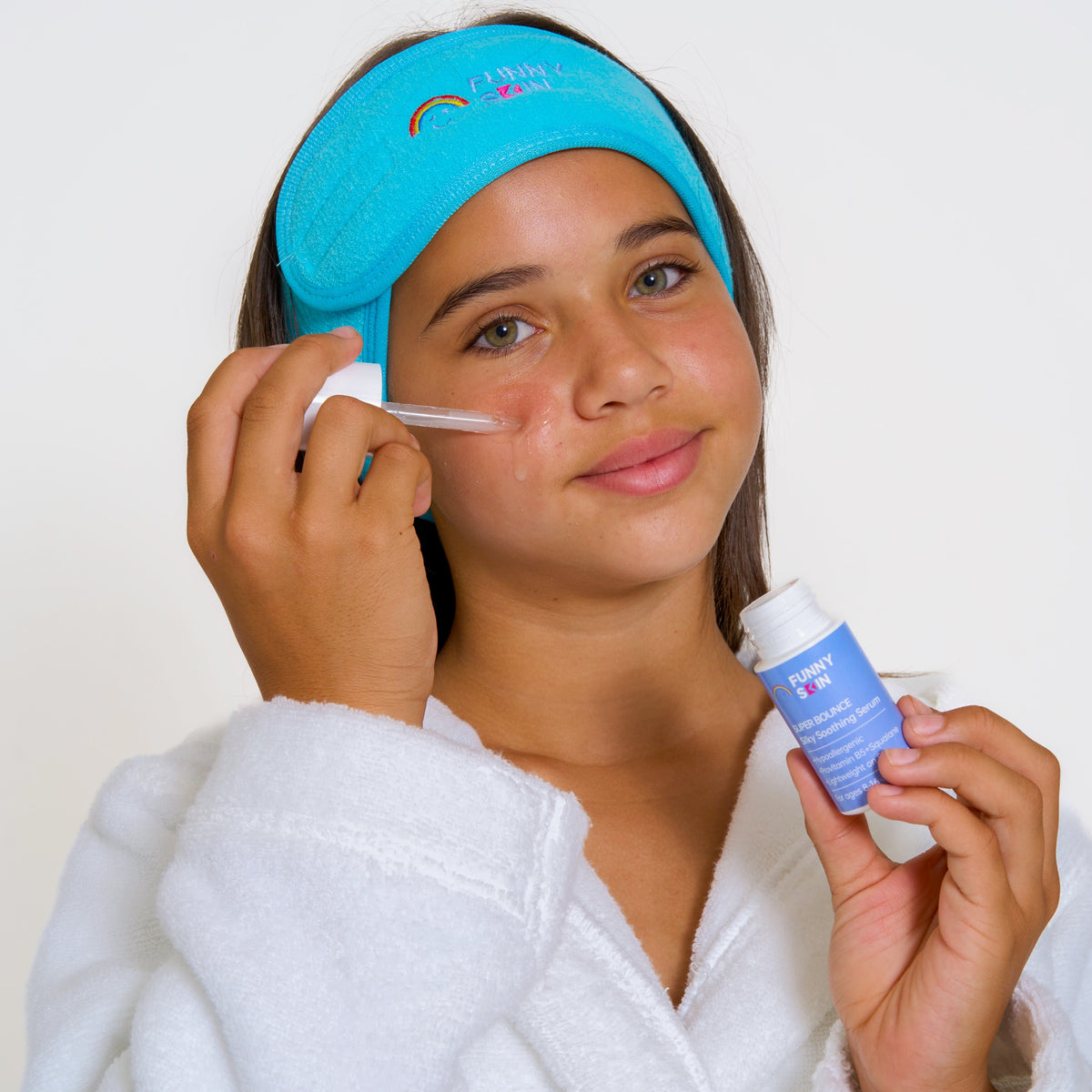 A young girl with a blue funny skin headband over her head, applying funny skin serum to her cheek