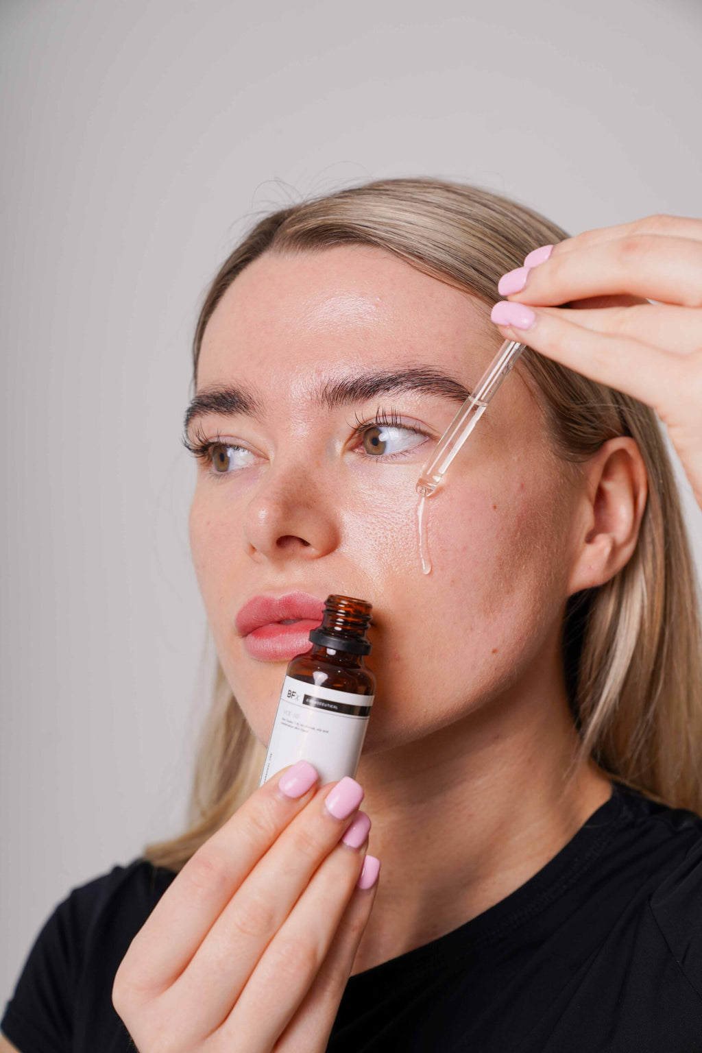 A woman applies a skincare serum from an amber dropper bottle using a glass pipette. She is holding the bottle and looking at the droplet as it drips onto her skin against a plain light gray background.