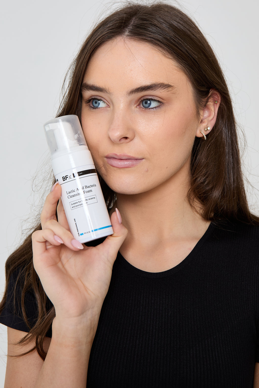 A woman with long brown hair holds a white bottle of Bossafce skincare cleansing foam next to her face against a light gray background.