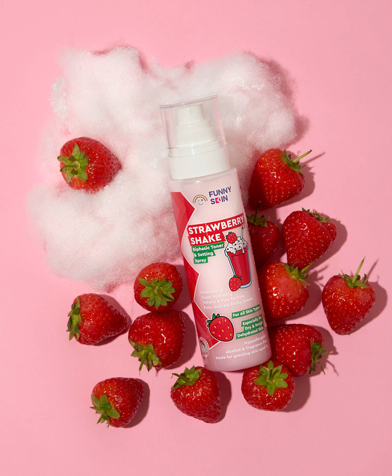 A bottle of Funny skin Strawberry Shake foam face spray, styled with fresh strawberries and white fluffy cotton candy on a solid pink background.