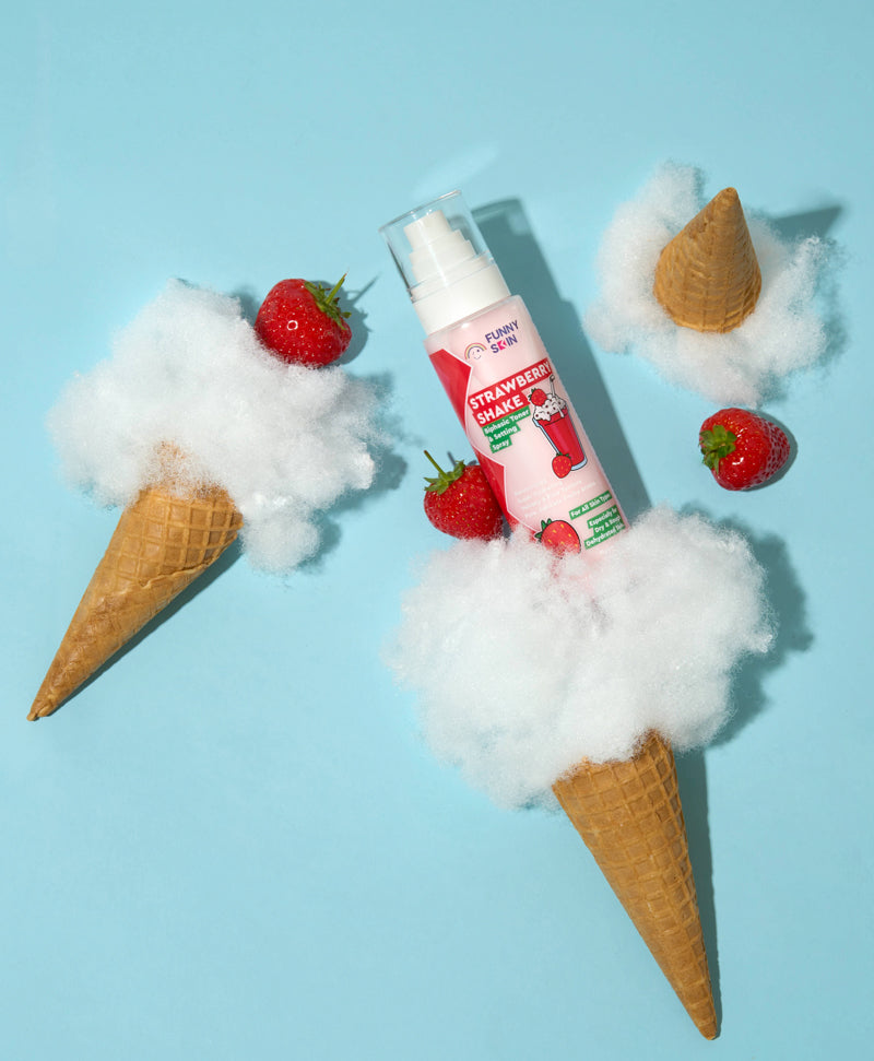 A tube of Funny Skin Brand Strawberry Shake face spray is styled in a flat-lay composition on a light blue background, surrounded by cotton candy, waffle cones, and fresh strawberries.