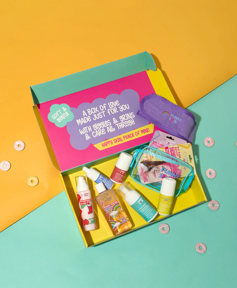 A colorful gift box of skincare products from Happy Skin Force of Mind, styled on a split yellow and mint green background. The open box contains various skincare items like body wash, hand cream, and a purple lip balm.