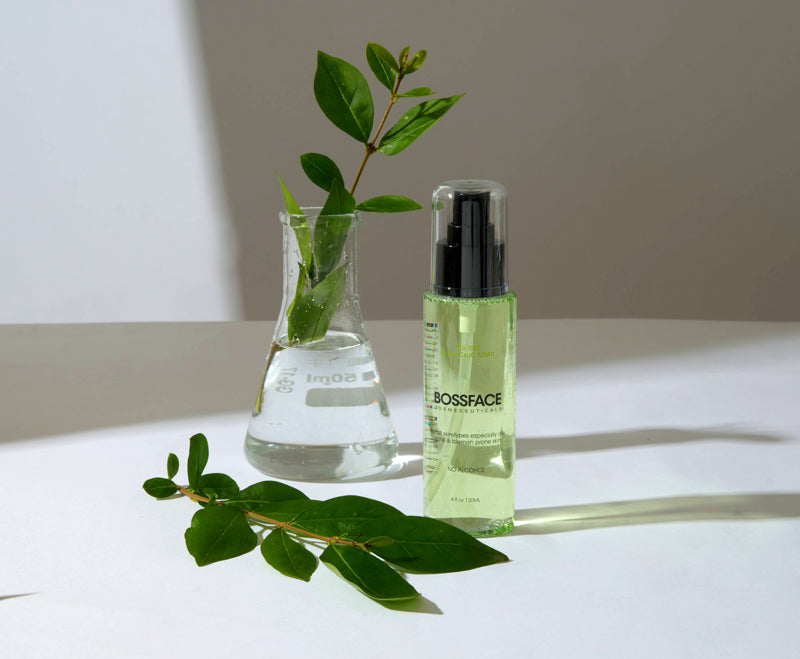 A clear bottle of BLOSSFACE serum with a black pump, filled with pale yellow-green liquid, standing on a white surface beside a small clear beaker with green leaves and a loose leafy branch in front, against a warm off-white background.