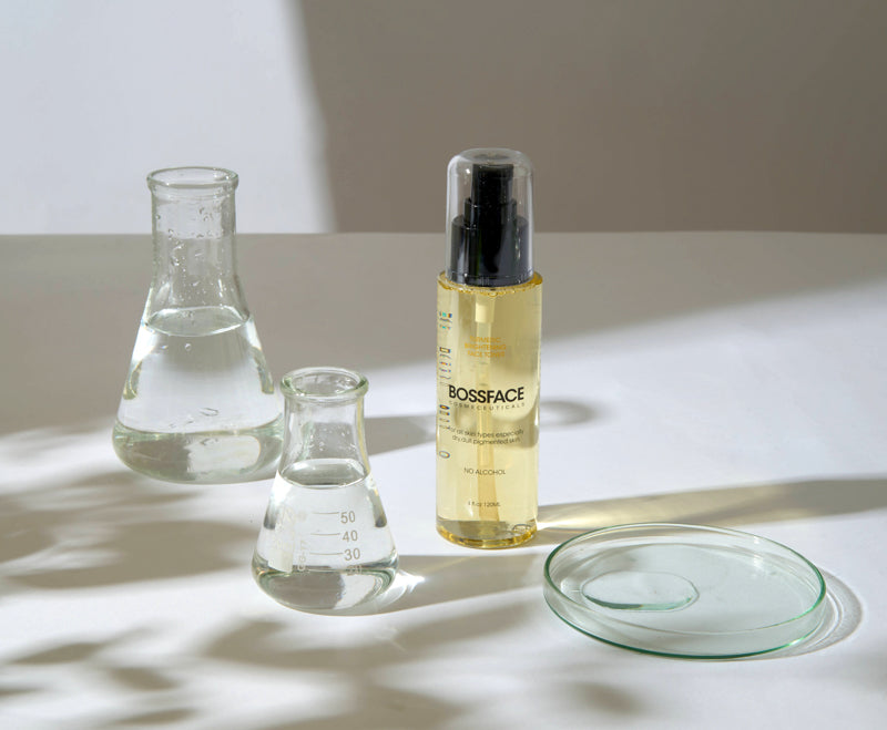 A bottle of BOSSFACE Turmeric Toner with a clear cap, standing next to two glass beakers and a glass lid on a white surface with dramatic shadows.