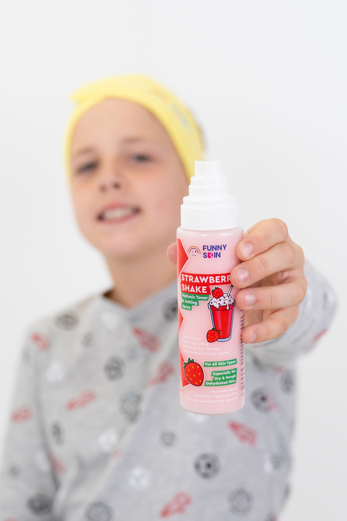 A child wearing a yellow headband and a gray patterned pajama shirt holds up a pink bottle of Funny Skin Strawberry Shake against a plain white background.