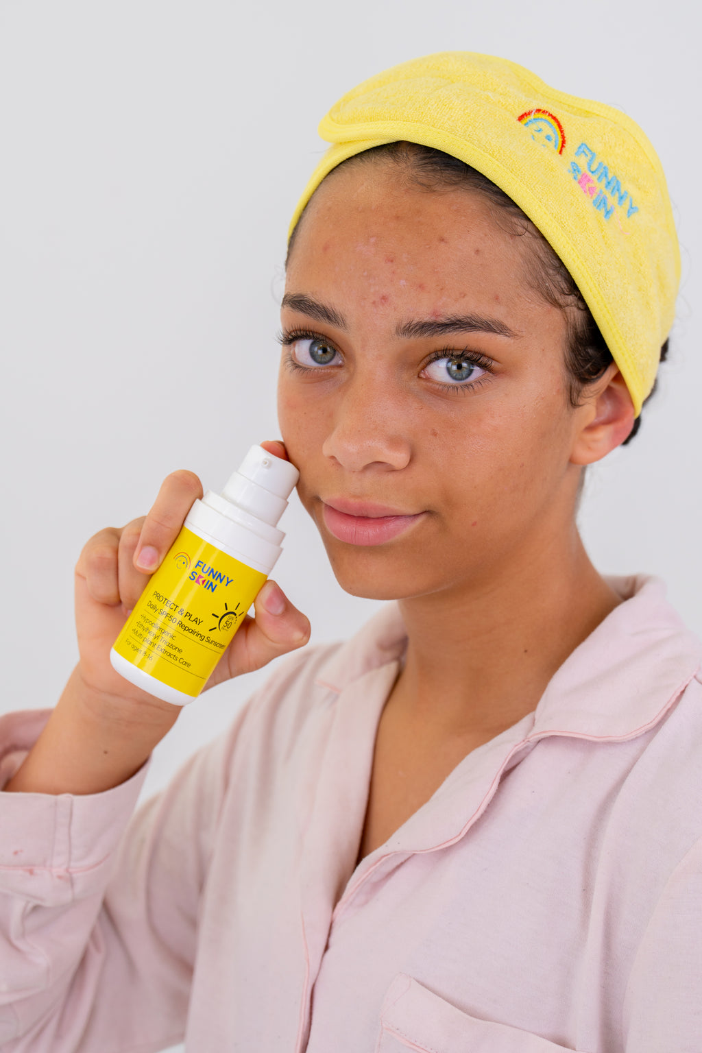 A young girl wearing a yellow towel cap and light pink pyjamas holds a yellow funny skin protect and play sunscreen bottle to her face against a plain light gray background.