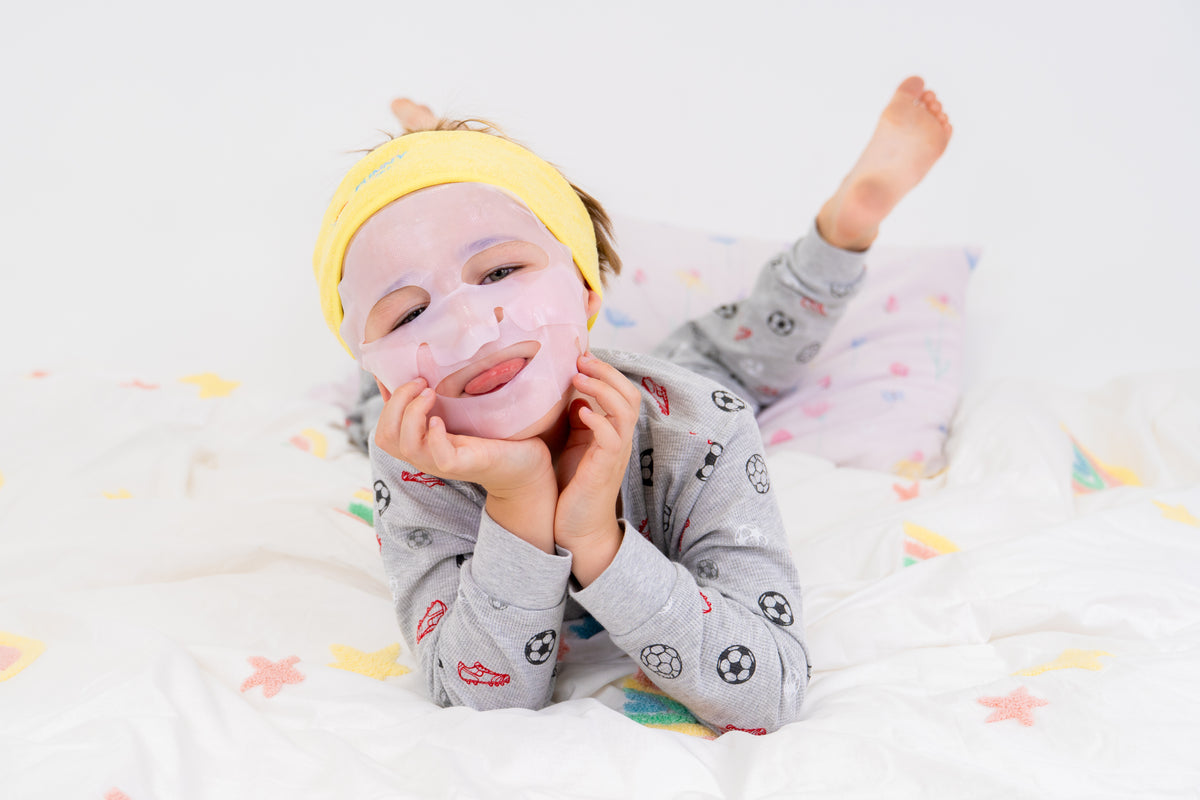 A toddler lying on a white bed wearing a gray long-sleeve pajama shirt with soccer ball prints, a yellow headband, and a pink face mask.