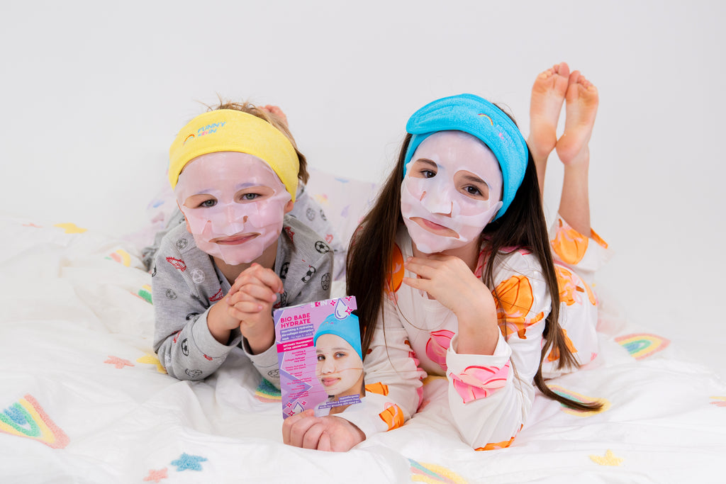 Two children lying on a white bed with star and rainbow prints, wearing sheet masks and headbands, with one child holding a packaged skincare product.