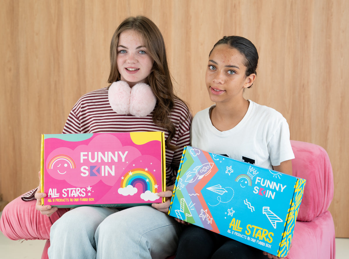 Two women sitting on a pink armchair holding colourful Funny Skin all stars boxes, with a light wood-paneled wall in the background.