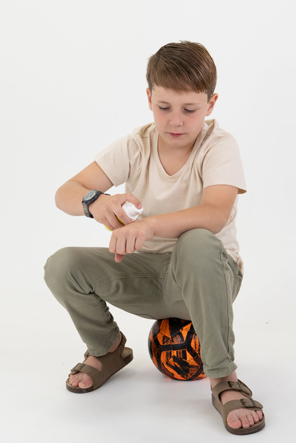 A young boy wearing a beige t-shirt, olive green pants, and brown sandals squats on a black and orange soccer ball while applying sunscreen from a white bottle to his arm against a white studio background.