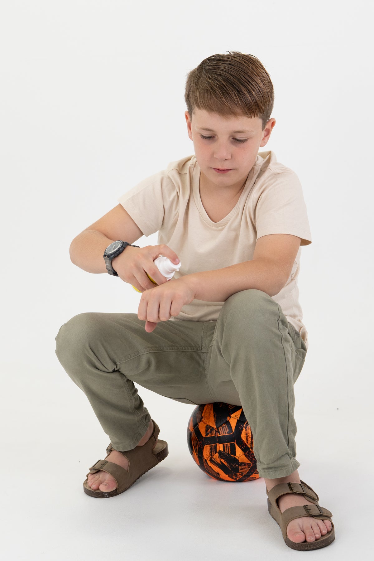 A young boy wearing a beige t-shirt, olive green pants, and brown sandals squats on a black and orange soccer ball while applying sunscreen from a white bottle to his arm against a white studio background.