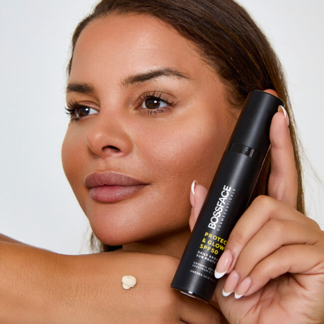 Woman holding Protect and Glow skincare product next to her cheek with cream on the back of her hand