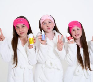 three young girls in bathrobes wearing funny skin headbands. All three girls are holding funny skin products