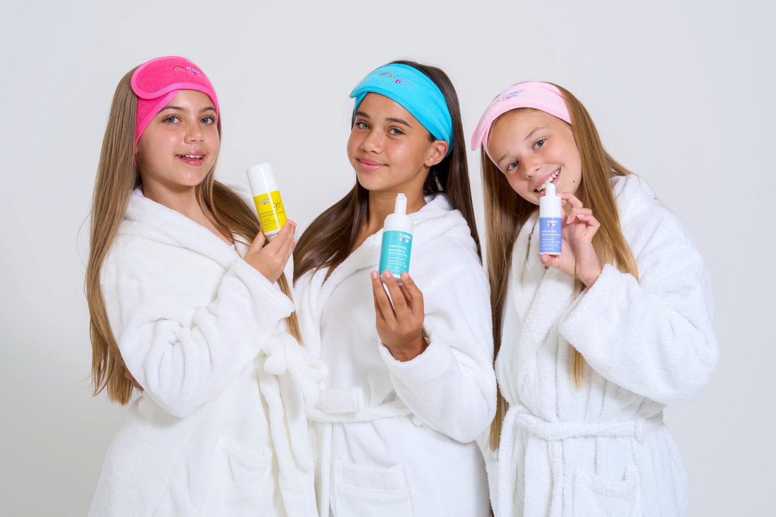 three tween girls in bathrobes wearing funny skin headbands. All three girls are holding funny skin products